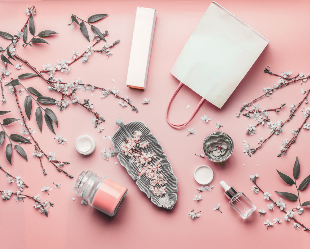 Cosmetic concept. Various facial products and paper shopping bag on pastel pink background with cherry blossom and leaves, top view, frame. Copy space for your design. Beauty blog layout. Flat lay