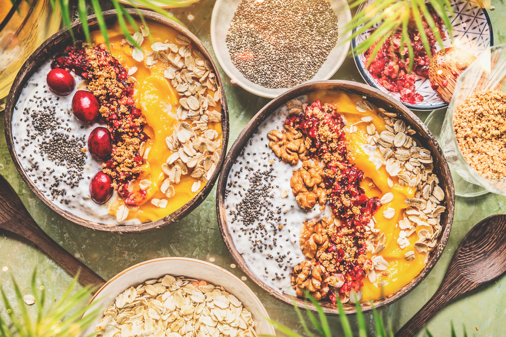 Smoothie mango bowls with chia seeds yogurt pudding and cranberries, nuts, oatmeal topping in coconut shells with spoon, top view. Healthy clean breakfast food