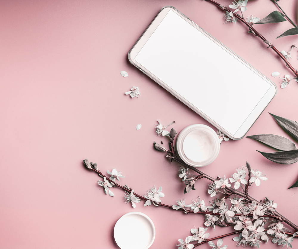 Smartphone mock up on pastel pink desktop background with cosmetic and blossom branches, top view. Beauty blog and female business concept