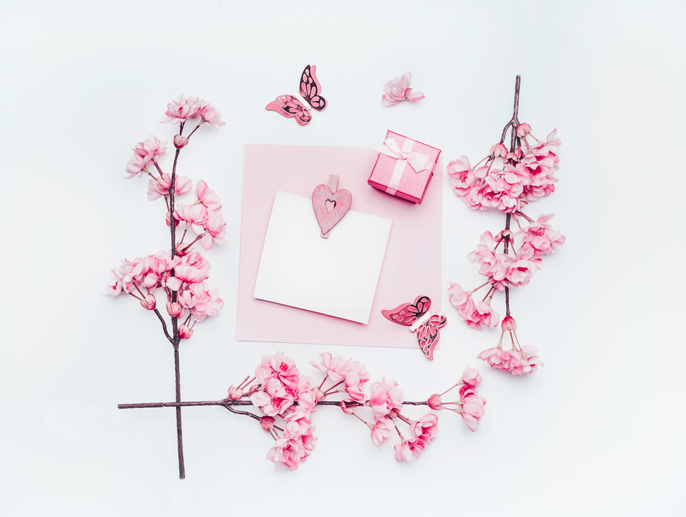 Blank greeting card mock up with pastel pink spring blossom flowers, gift box and hearts on white background, top view. Mothers day, wedding or birthday concept. Copy space for your design.