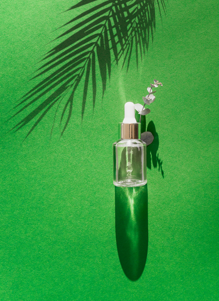 Transparent cosmetic bottle with pipette standing on green table with palm leaves shadow. Facial skin care concept. Natural vegan cosmetic. Serum or skin essential oil. Modern beauty trend.