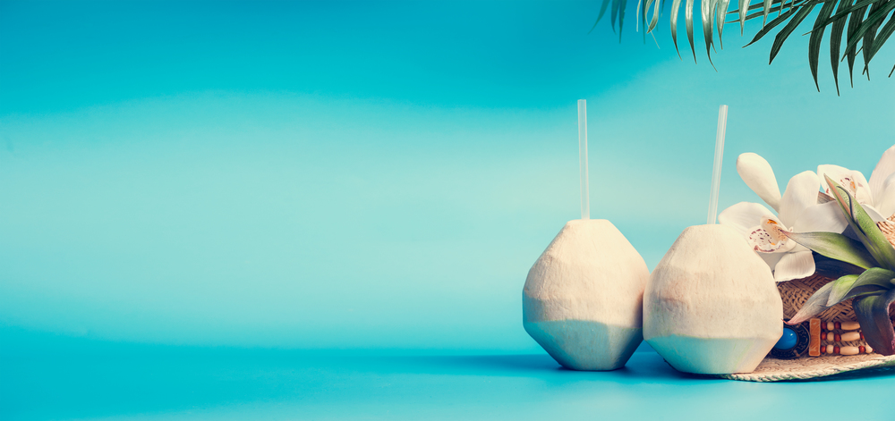 Summer tropical vacation background banner with fresh tropical coconut cocktails , drinking straws and palm leaves and flowers standing on blue turquoise background. Travel and holiday concept