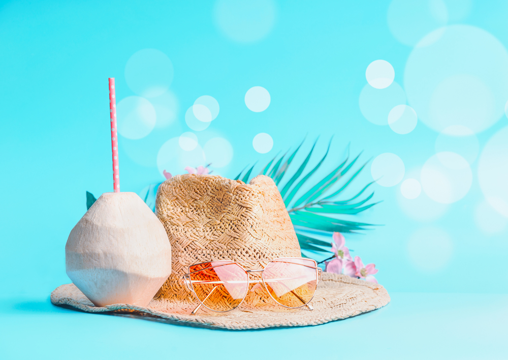 Womans beach accessories with fresh coconut, drinking straws and tropical leaves and flowers , sunglasses and straw hat on sunny blue background with bokeh. Summer holiday concept, banner.