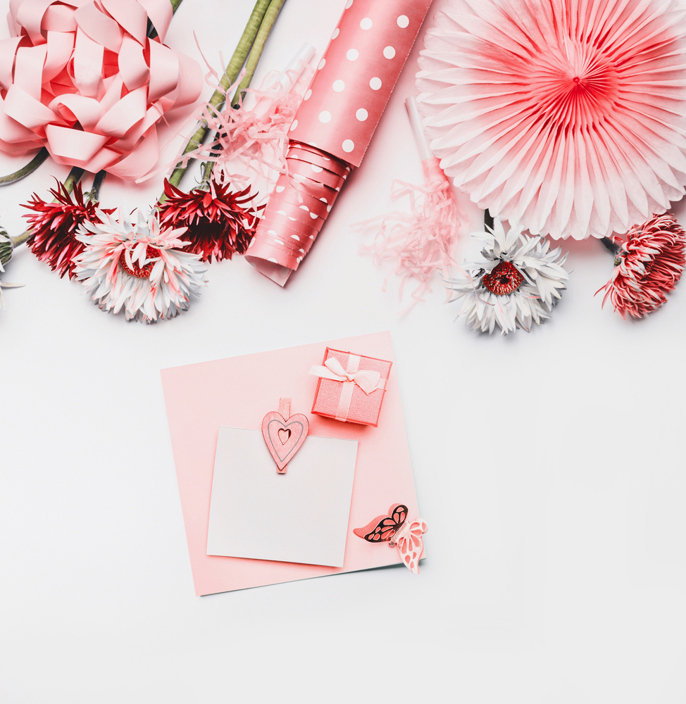 Female still life greeting objects in coral color on white background with greeting card mock up. Ribbon , paper party fan, flowers and dotted wrapping paper on white background.  Flat lay, top view.