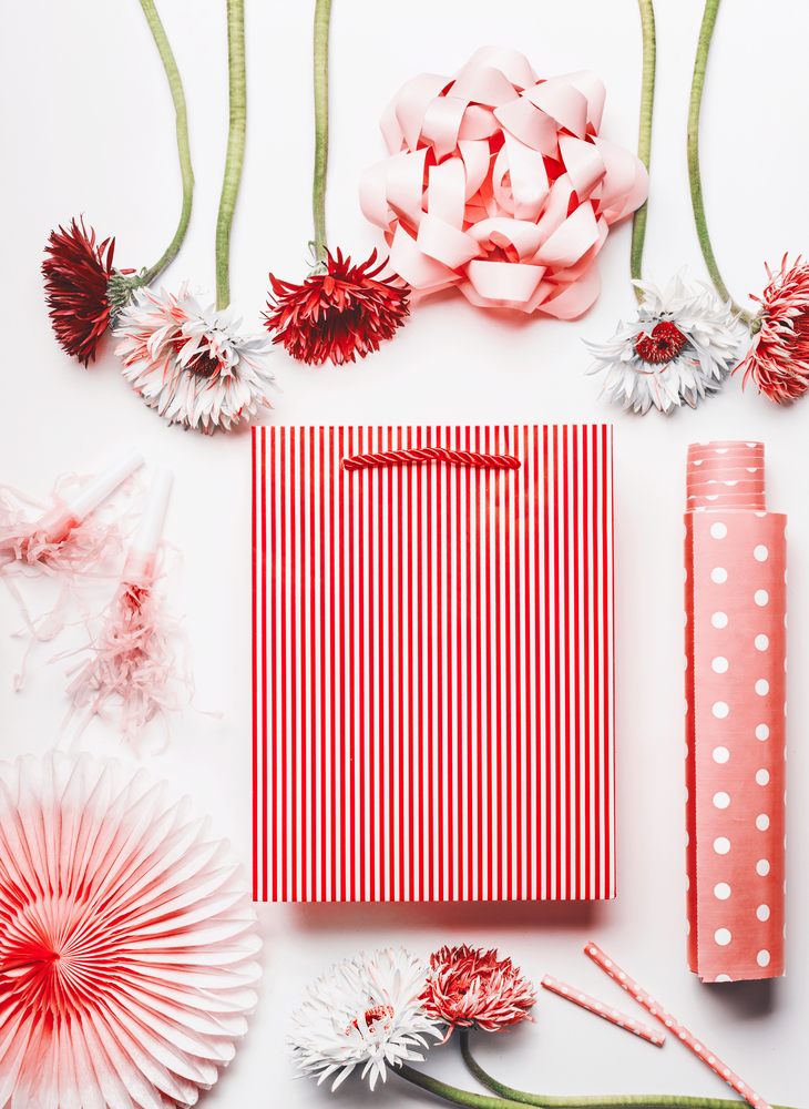Female greeting objects still life in coral color on white background. Striped shopping bag ,ribbon , paper party fan, flowers and dotted wrapping paper on white background.  Flat lay, top view.