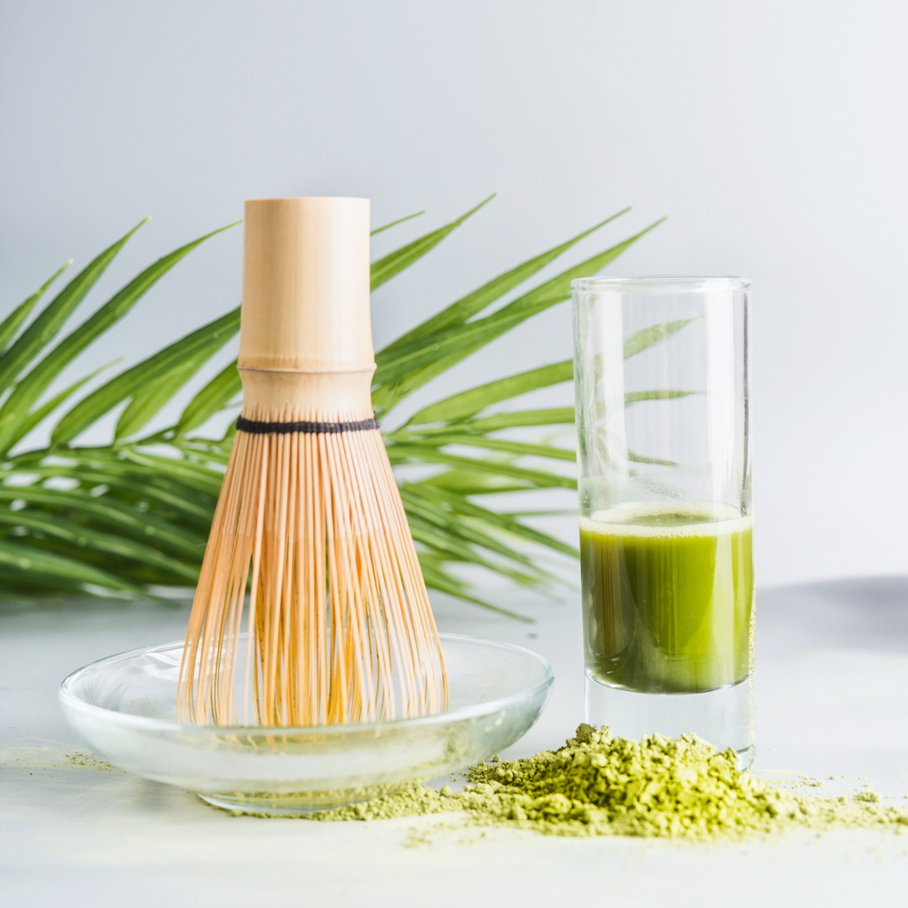 Close up of matcha espresso in glass with whisk and matcha powder on table at light background, front view with copy space. Clean, detox beverage, healthy dairy food concept. Antioxidant boost drink