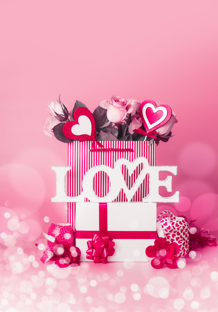 Valentines day concept. Gift box with roses bunch and hearts in shopping bag, word Love and ribbons standing at red pink background with bokeh, front view