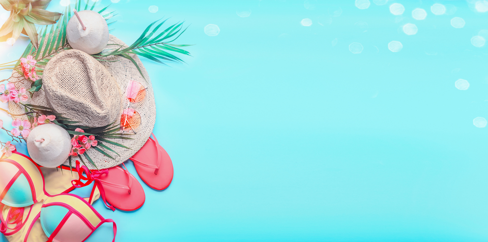 Top view of woman&rsquo;s summer beach accessories: bikini,  flip flops, sunglasses, straw hat, creole earrings, with palm leaves and tropical flowers on turquoise blue background, banner with copy space