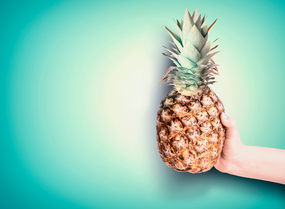 Hand holding pineapple at turquoise background. Summer tropical fruits concept. Copy space for your design