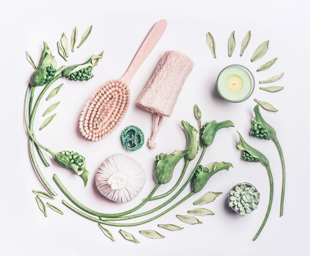 Various anti-cellulite equipment for dry massage at home on white background wit green leaves and flowers, top view. Dry brushing and skin pilling. Beauty and body care concept