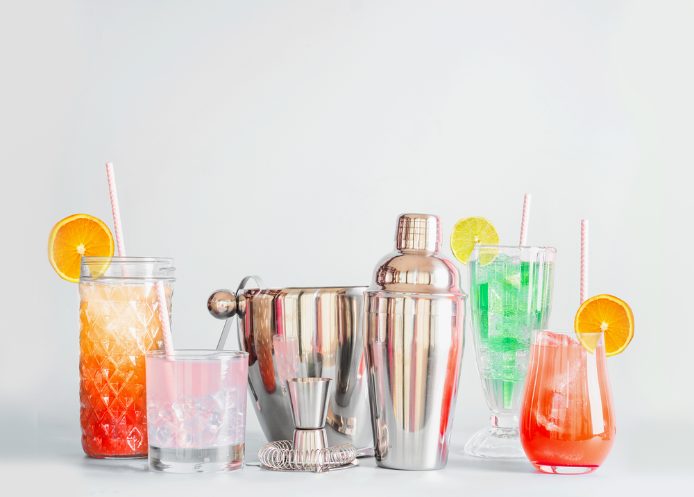 Colorful summer long drinks and cocktails bar tools in various glasses with paper drinking straws and citrus fruits at light background with copy space