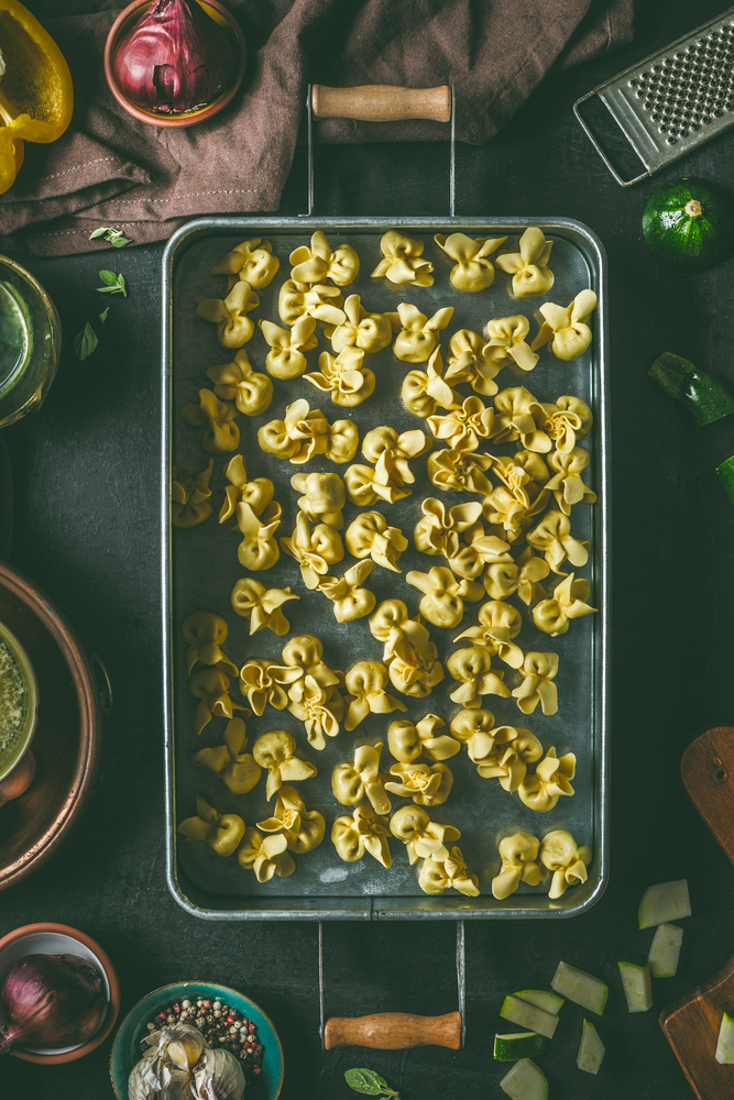 Homemade raw vegetarian tortellini pasta in metal tray on dark rustic kitchen table background with ingredients and utensils, top view. Cooking preparation. Italian food