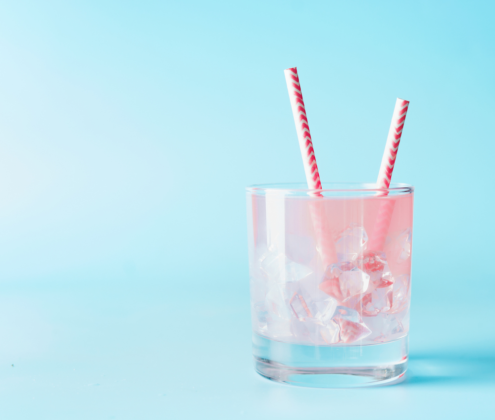 Pink iced refreshment drink in glass with paper drink straw on blue background. Copy space. Summer pastel pink cocktail.