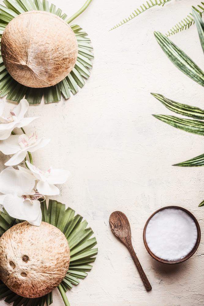 Wooden bowl with coconut oil and spoon on light background with coconuts, tropical leaves and flowers. Natural organic  concept. Top view.  Vegan coconut butter. Vegan ingredients. Healthy lifestyle