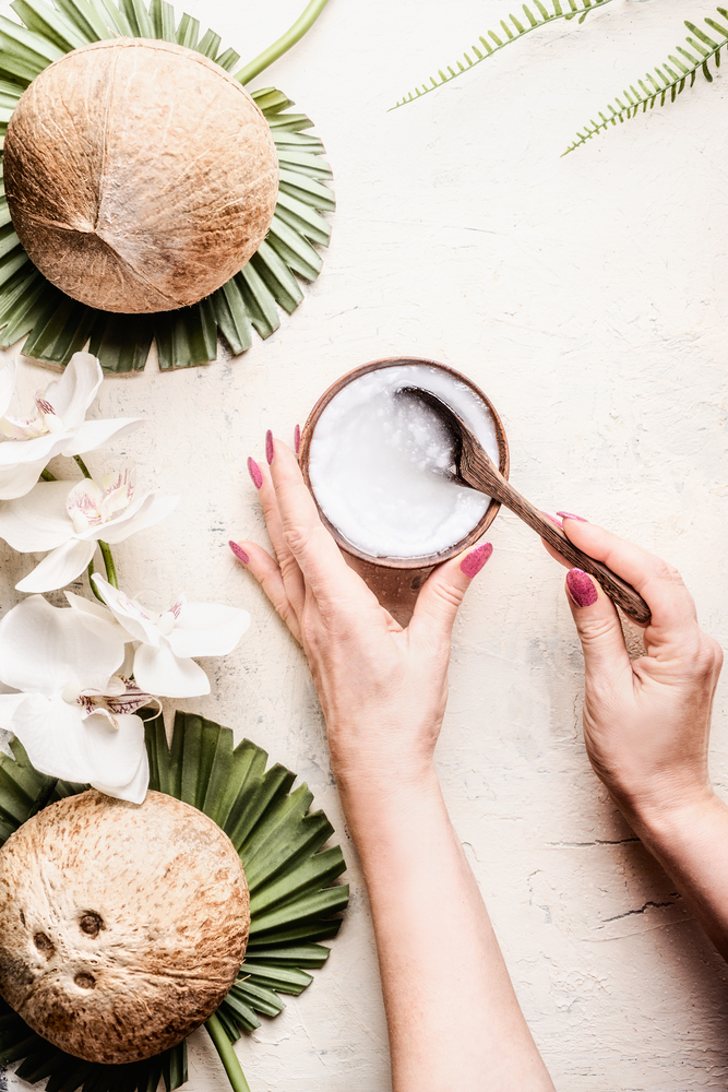 Female hands holding bowl and spoonful of coconut oil light background with coconuts, tropical leaves and flowers. Natural organic cosmetic concept. Top view. Vegan butter