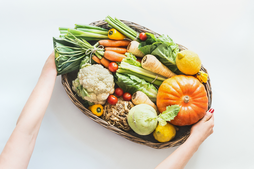 Female hand holding tray with various colorful organic farm vegetables from local markt on white desk background. Healthy food and clean seasonal eating concept. Top view. Clean seasonal eating