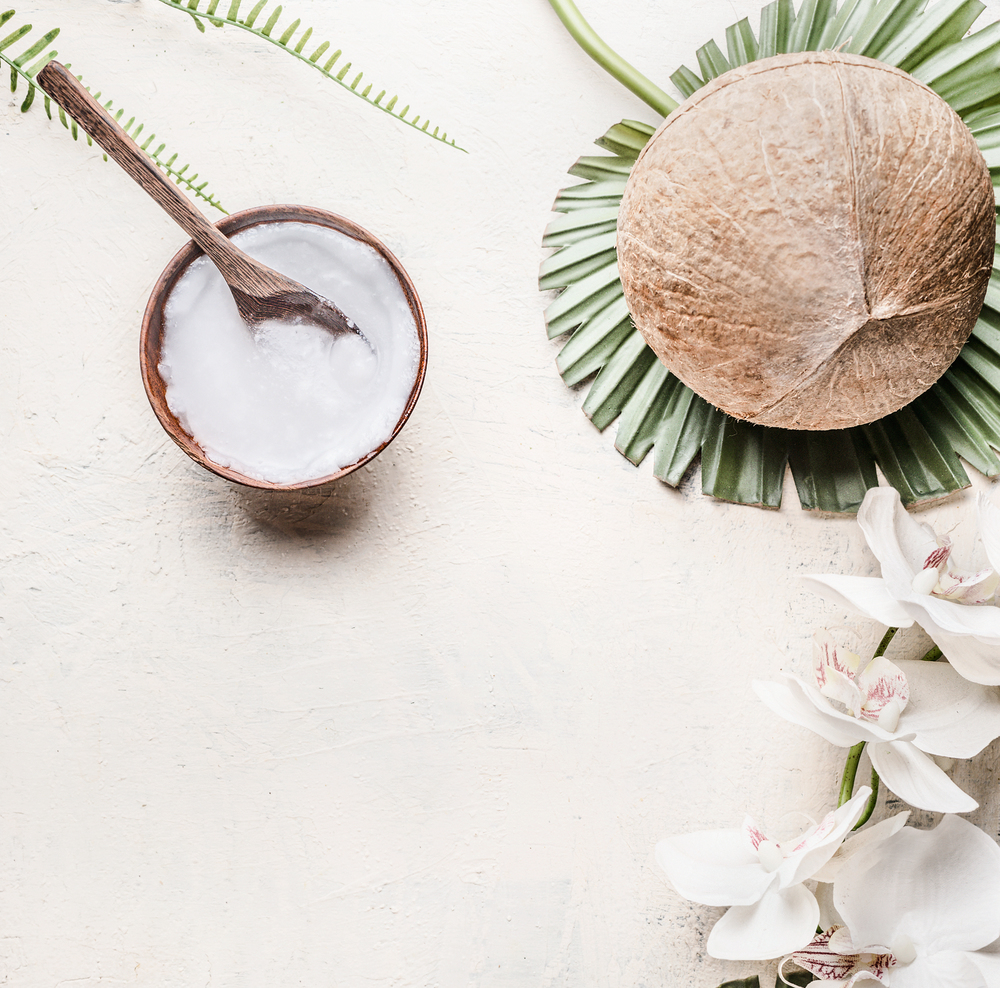 Coconut oil or butter in wooden bowl with spoon and whole coconut on tropical leaves and flowers, top view . Copy space . Healthy plant based fat source. Vegan food and cosmetic