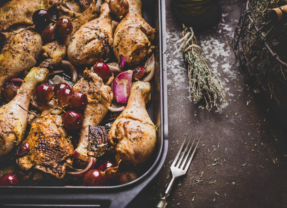 Close up of roasted chicken drumsticks with red onion and grapes on dark background.