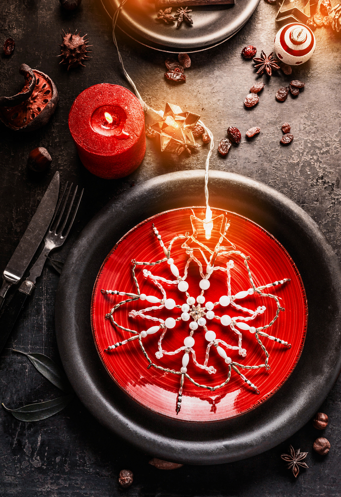 Christmas dinner table setting with red and black plates, decorated with snowflakes, burning candles and fairy lights on dark background. Top view