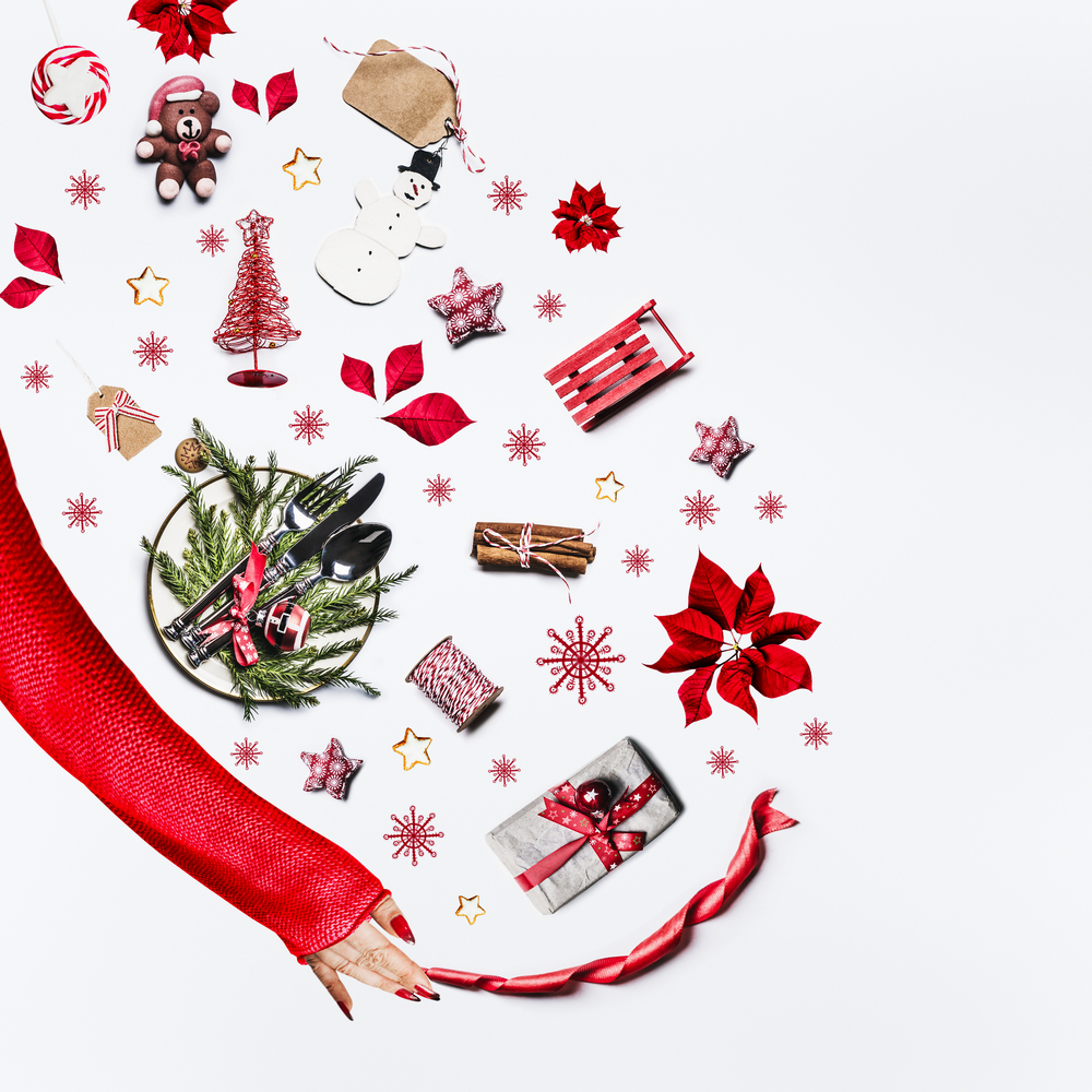 Woman hand in red sweater and various Christmas objects on white background, top view. Flat lay of Christmas table setting, holiday decoration and gift boxes with female hand. Fla lay.