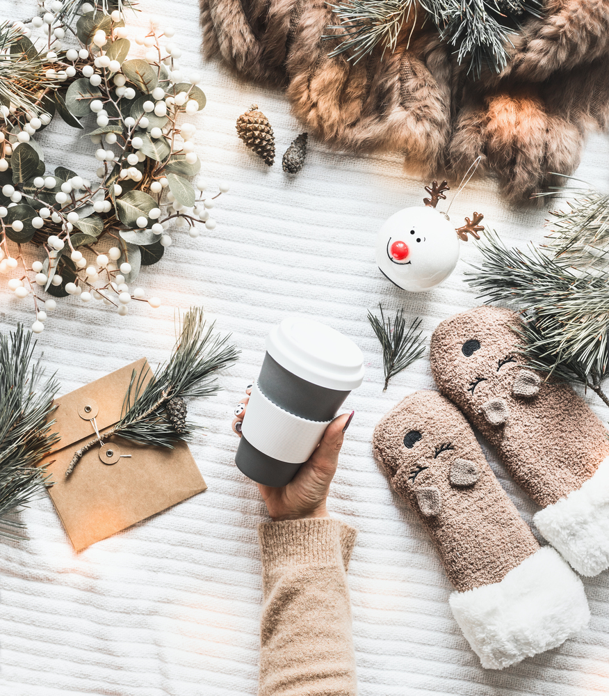 Female hand holding reusable eco  friendly coffee cup  on white knitted blanket with cedar branches, winter wreath, funny socks and craft paper envelope. Top view. Flat lay. Zero waste Christmas