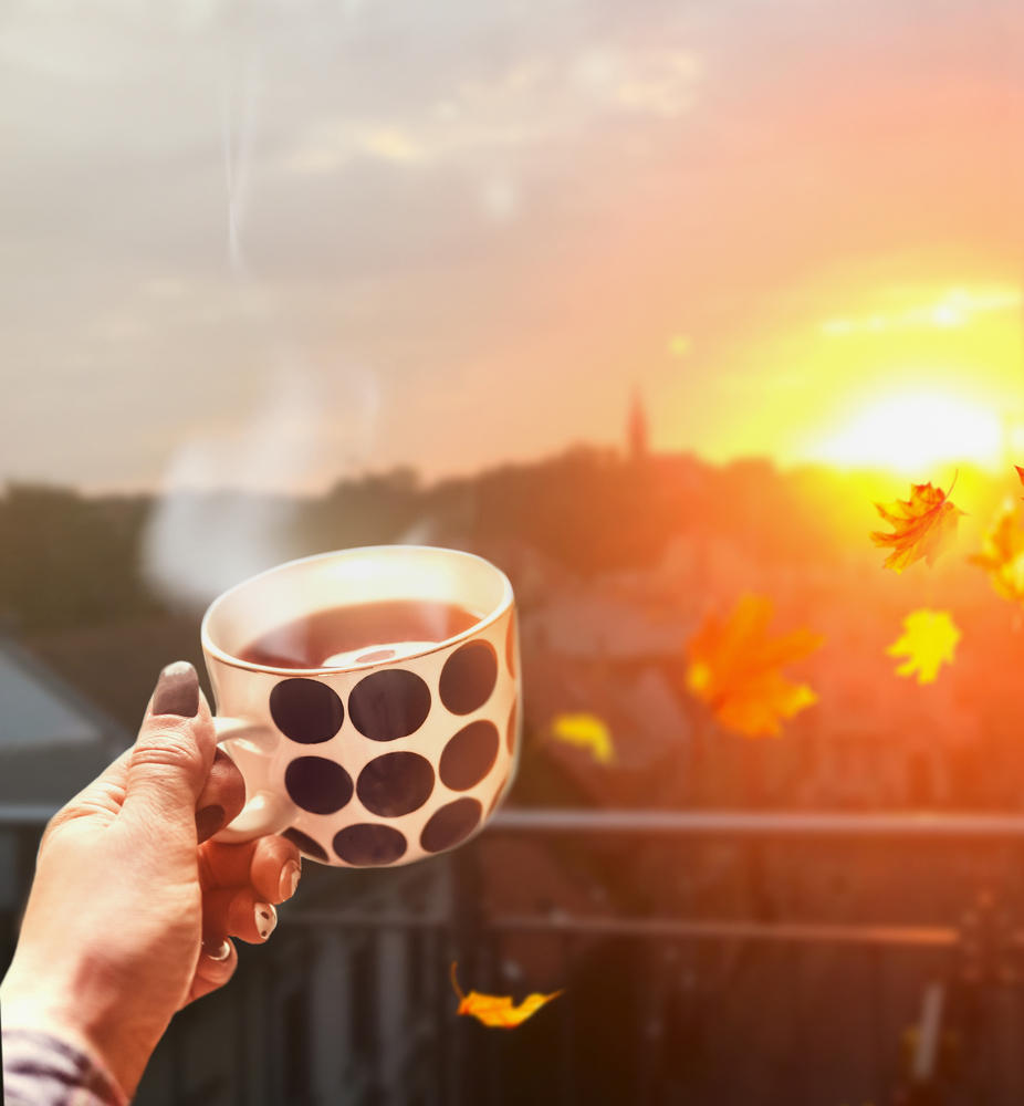 Woman hand holding cup with hot drink and steam at autumn city background with flying leaves and sunset light