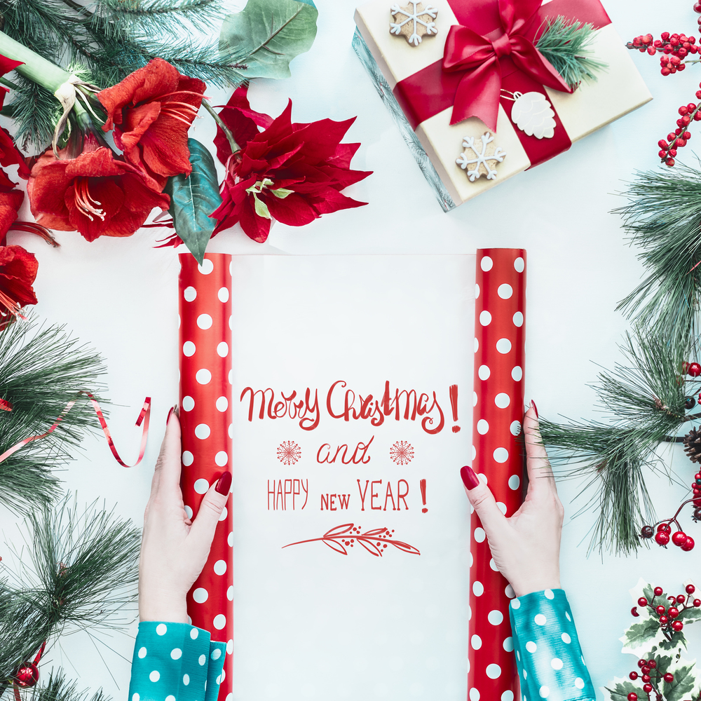 Female hands in blue blouse holding red polka dot wrapping paper with Merry Christmas and Happy New Year text on white desk with Christmas flowers , gift box and fir branches, top view. Flat lay