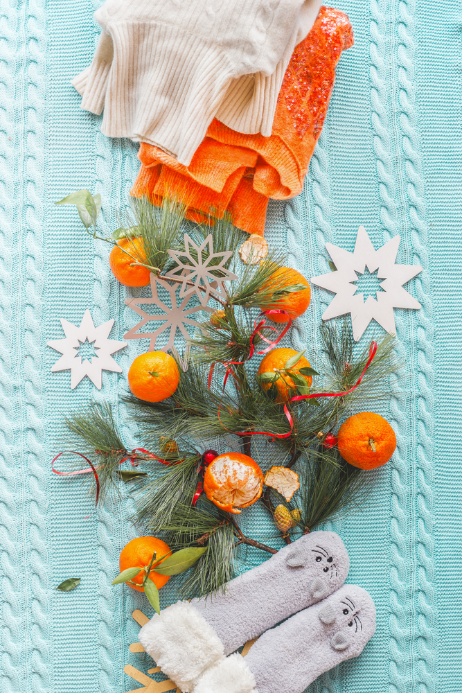 Winter mood composing with tangerines, orange sweater, funny socks and fir branches on blue knitted blanket with snowflakes. Top view. Copy space. Flat lay