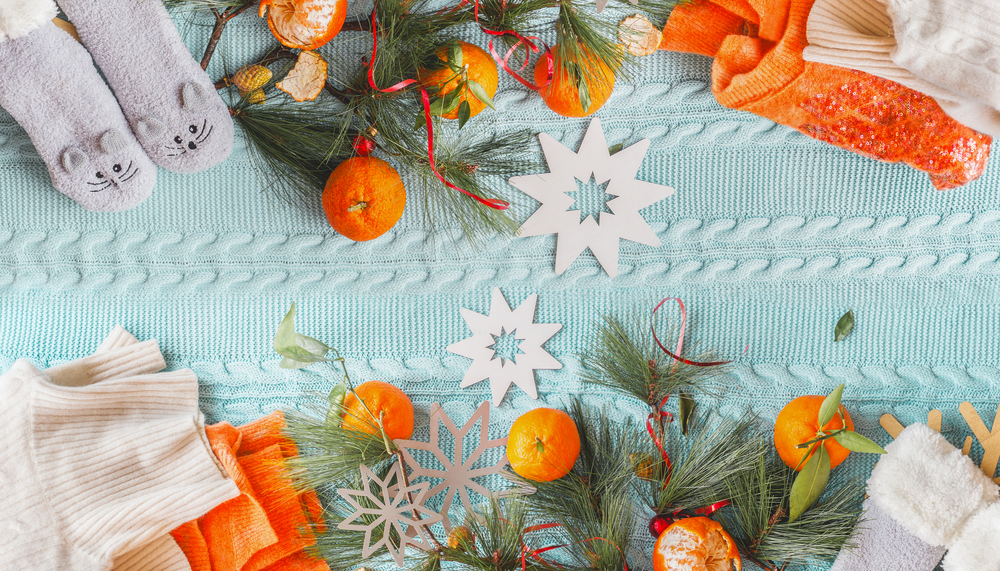 Cozy winter mood flat lay with tangerines, orange sweater, funny socks and fir branches on blue knitted blanket with snowflakes. Top view. Copy space.