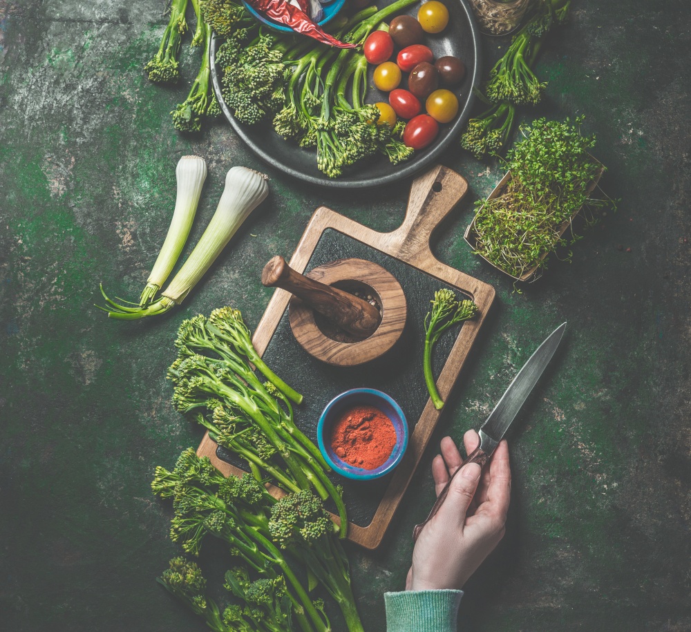 Cooking preparation of wild broccoli. Women hand holding knife on dark rustic background with cutting board and ingredients. Top view. Healthy food