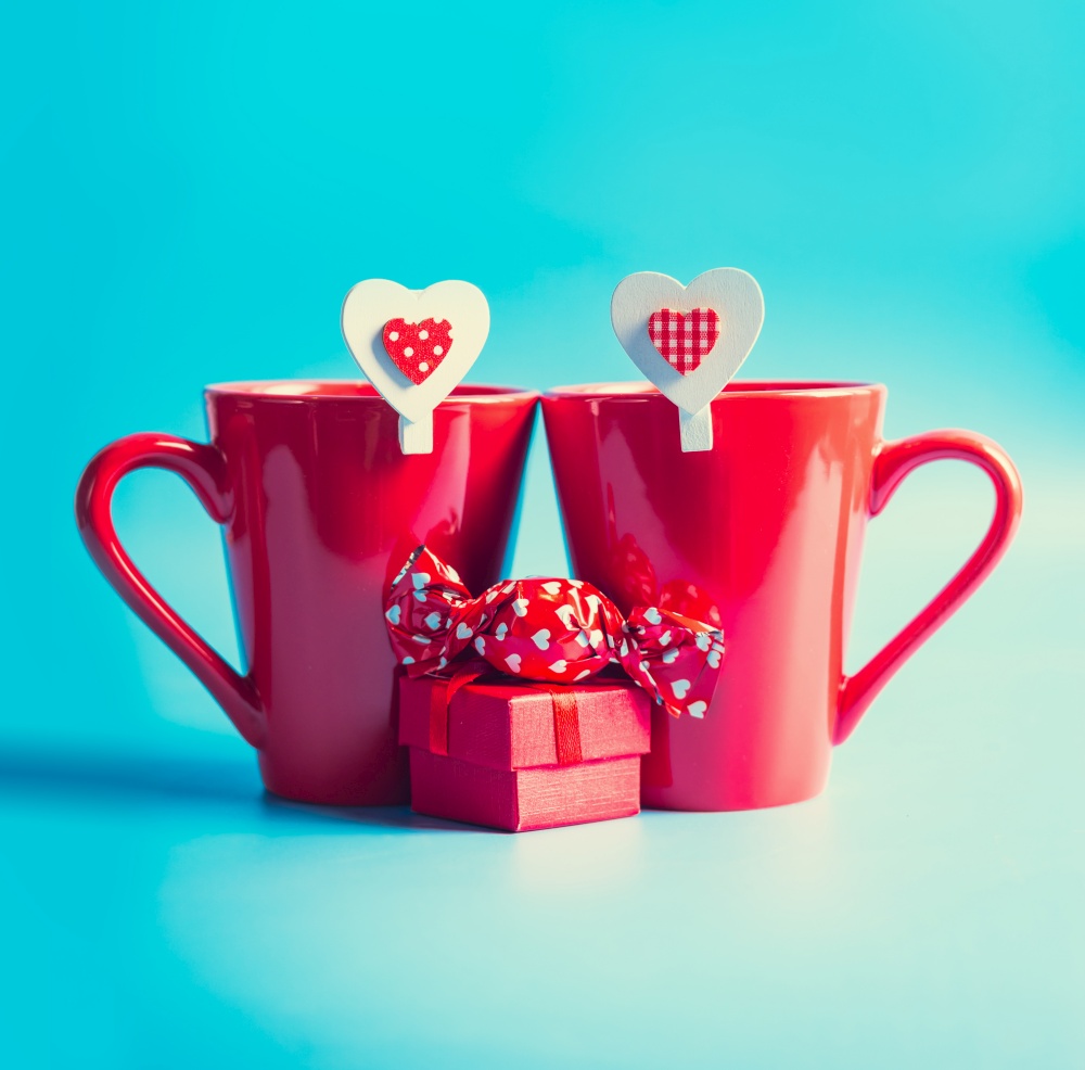 Two red mugs with hearts, candies and presents on blue background.