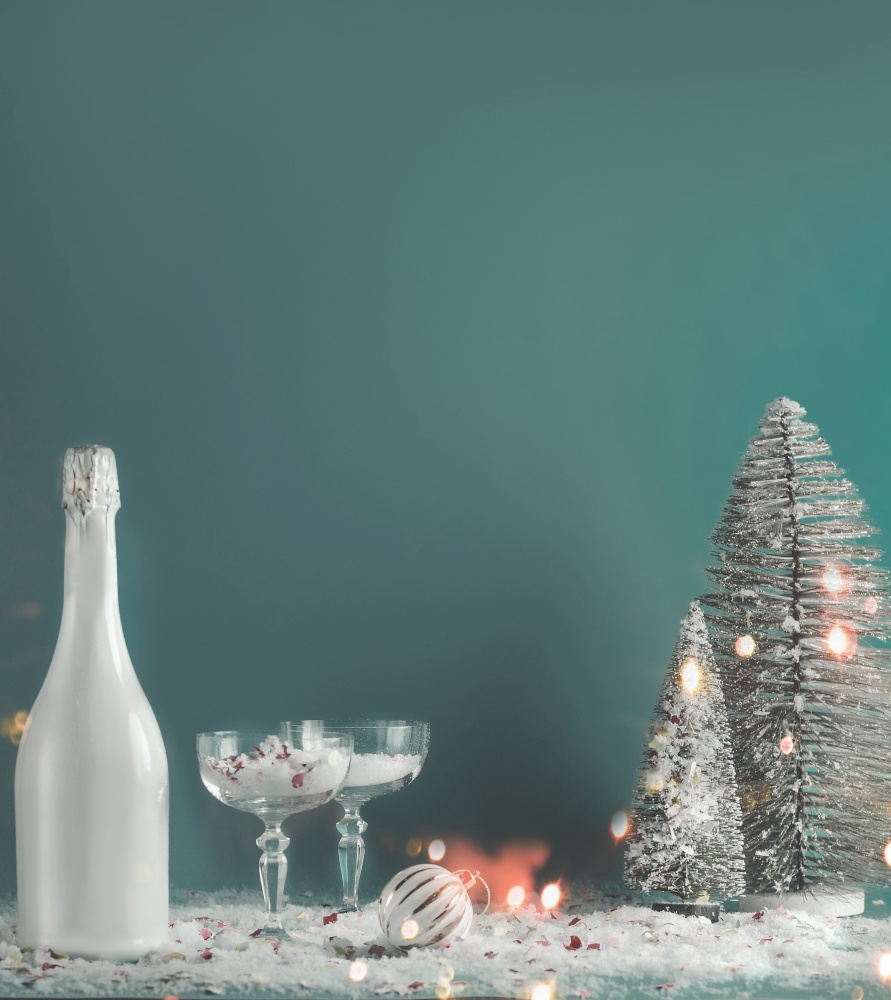 Christmas and New Year background with bottle of champagne , fancy glasses with snow, Christmas baubles, fir trees and fairy light bokeh. Holiday still life