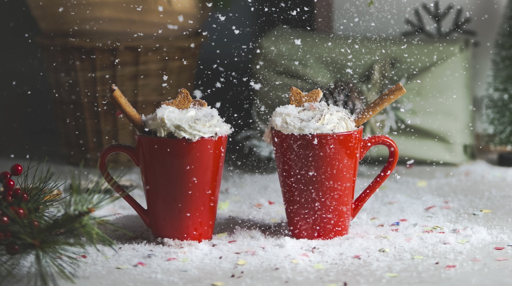 Two red mugs with Christmas drink hot punch or hot chocolate with whipped cream, cinnamon sticks and CHristmas cookie on table with snow. Front view. Cozy still life