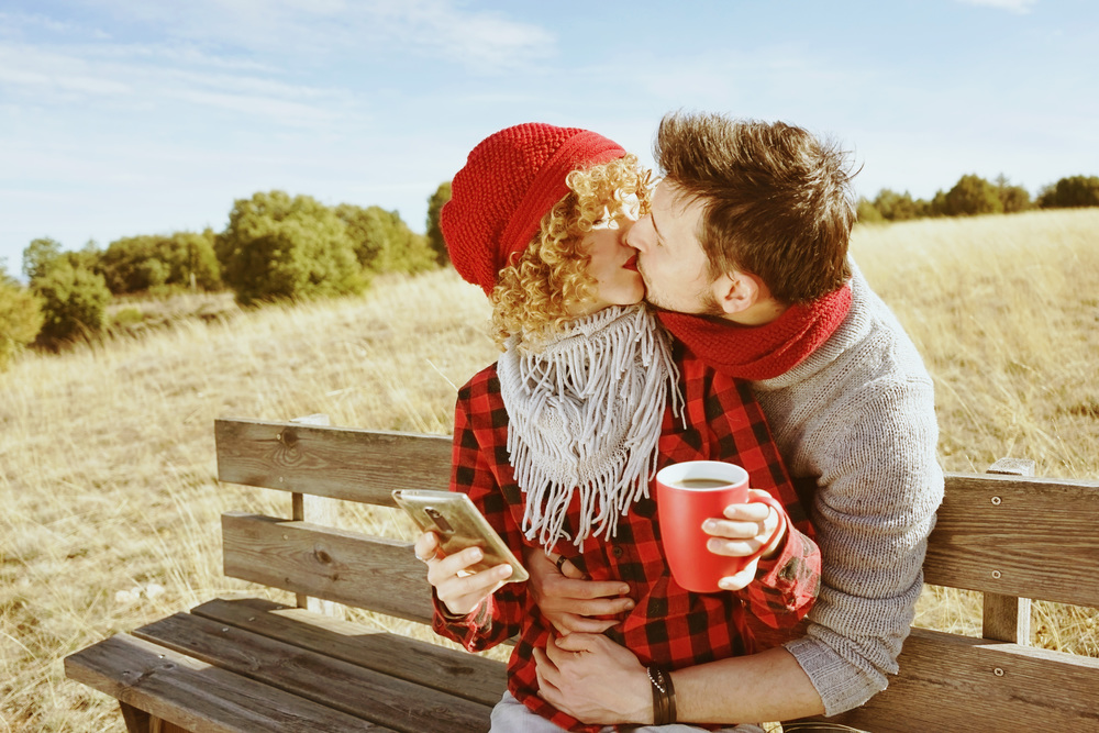 A couple of happy young millennials in love sunbathing on a wooden bench and taking a cup of coffee or tea while they are kissing and holding a smartphone with the backlight from autumn sun