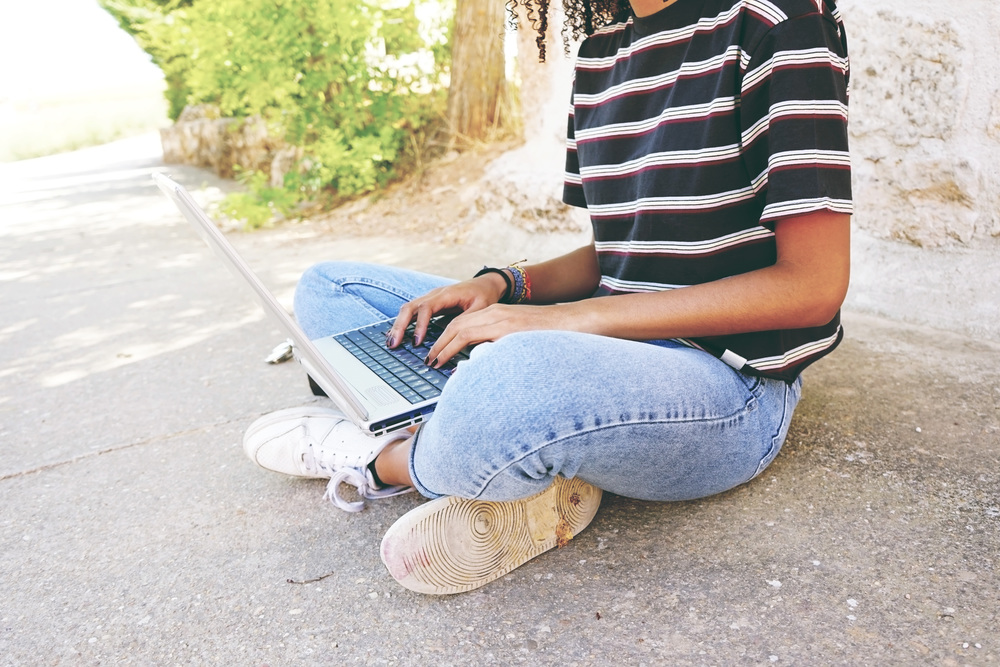 A young black woman with curly hair wearing jeans and a striped t-shirt, sitting on the ground and working or making homework