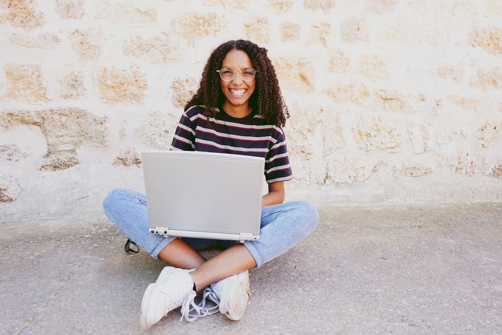 A portrait of happy smiling young black woman with curly hair wearing glasses, jeans and a striped t-shirt, sitting on the ground and working or making homework