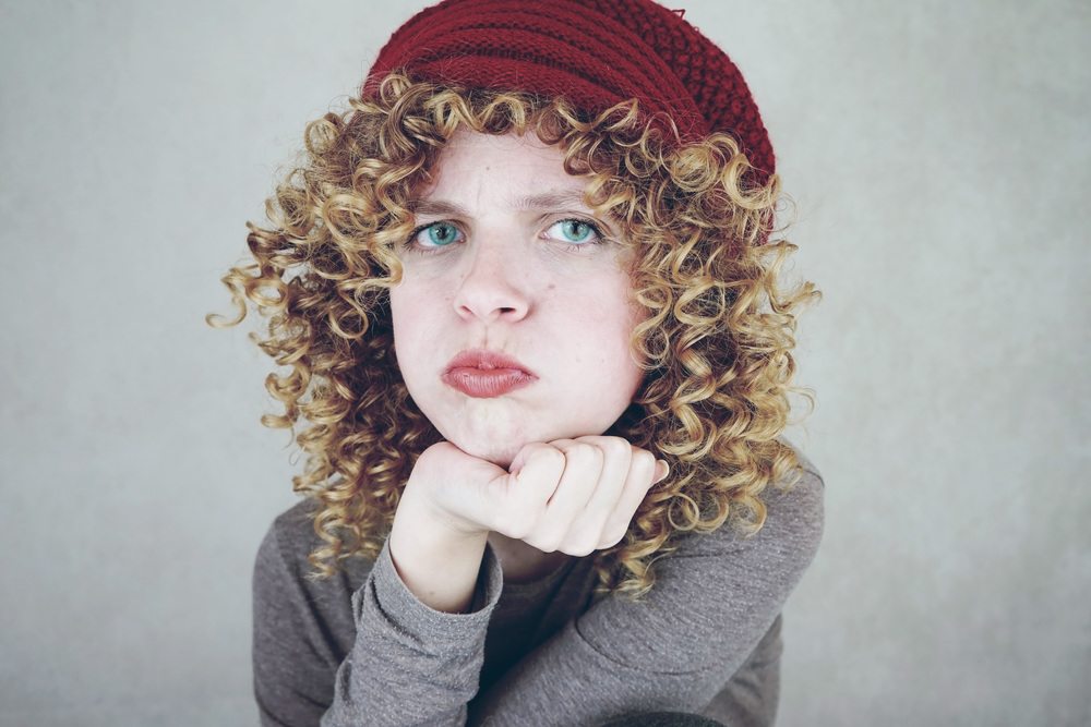 Close-up portrait of a beautiful and young funny bored or angry woman with blue eyes and curly blonde hair wearing a red woolen cap