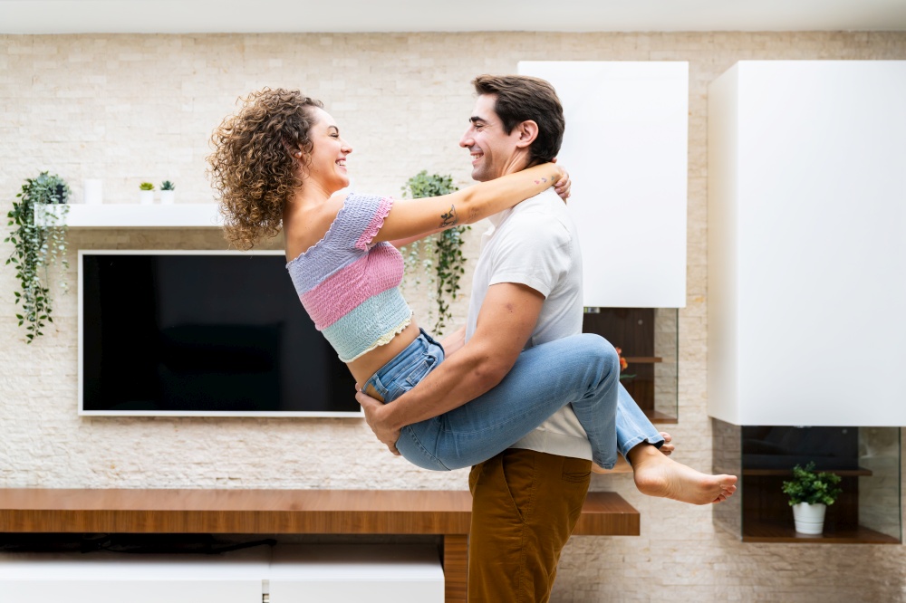 Side view of smiling young couple embracing while having romantic moment in modern apartment against LED TV and looking at each other happily. Cheerful young couple hugging at home