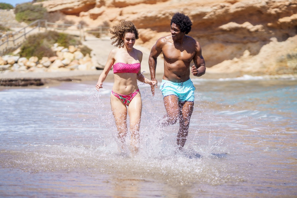 Full body of multiracial couple in swimwear running in splashing water and having fun while looking away and enjoying free time near seaside in summertime. Happy diverse couple jogging in sea water