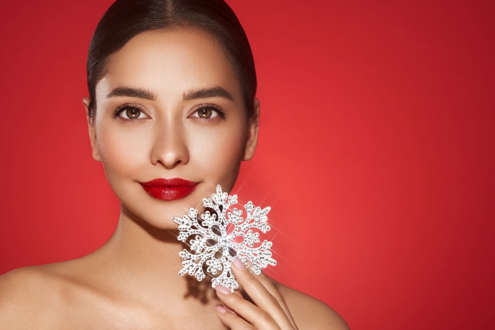 Beautiful young woman with red lipstick, shiny clean skin and face, showing glowing snowflake, near face and smiling, isolated on a red background. Christmas holidays concept