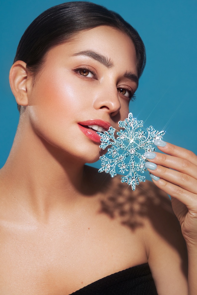 Beautiful young woman with red lipstick, shiny clean skin and face, showing glowing snowflake, near face and smiling, isolated on a blue background. Christmas holidays concept