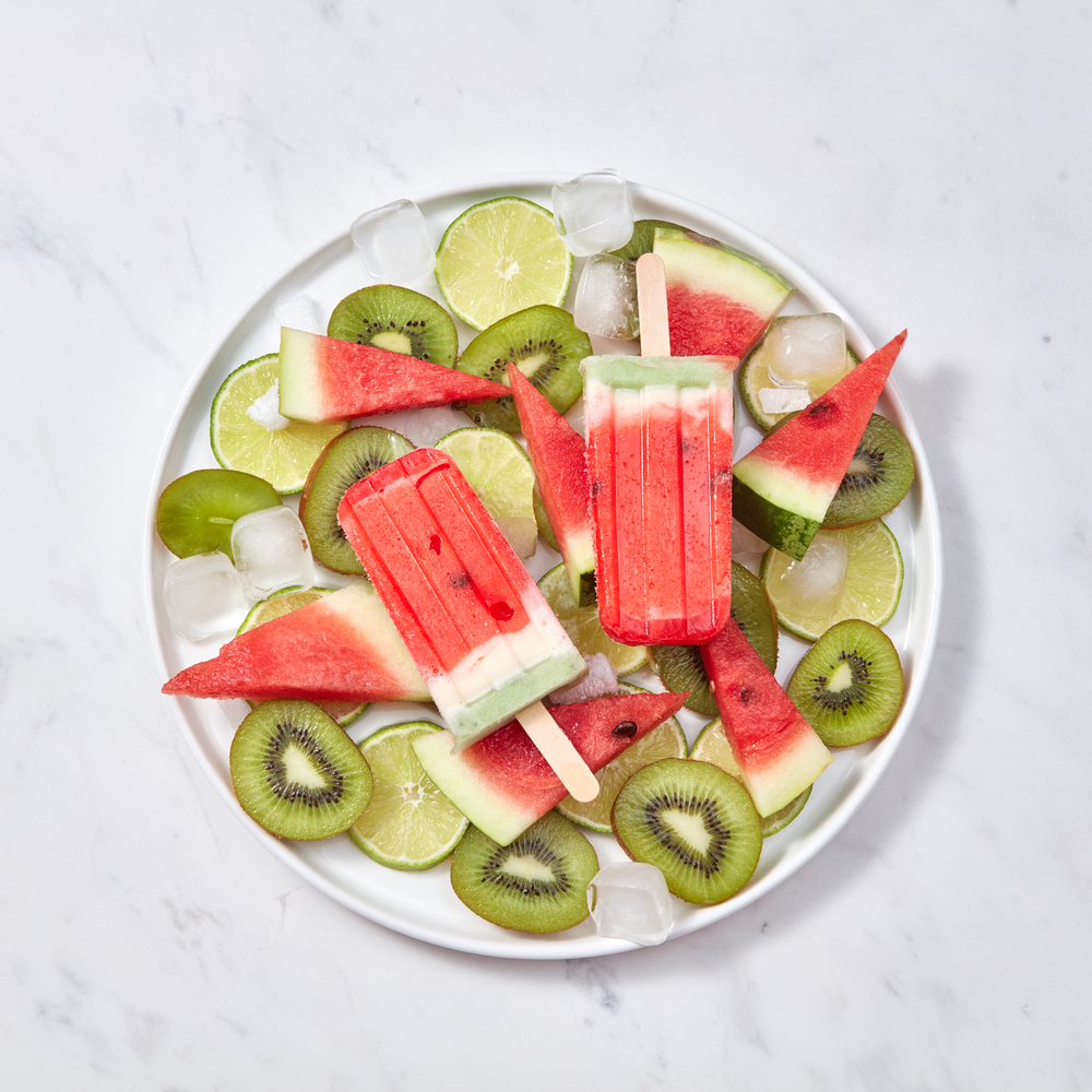 Colorful icy popsicle. Appetizing homemade watermelon ice cream in a plate with slices of lime, watermelon, kiwi and ice cubes on a gray marble table with a copy of the space. Flat lay. Fruit ice cream on a stick is presented in a plate with ice cubes and pieces of lime, watermelon and kiwi on a gray marble background. Space for text. Flat lay