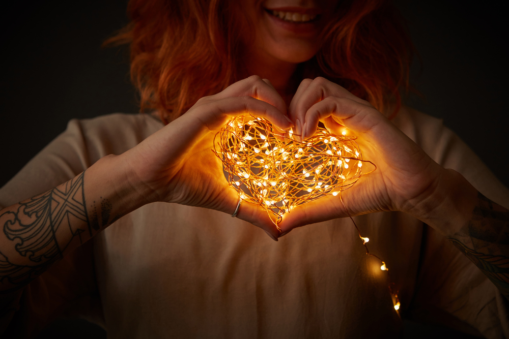 A smiling girl with a tattoo is holding in her hands a bright garland in the shape of a heart around a dark background. Happy Valentine&rsquo;s Day. Happy red-haired girl holding garland in the shape of heart on a dark background. Festive card
