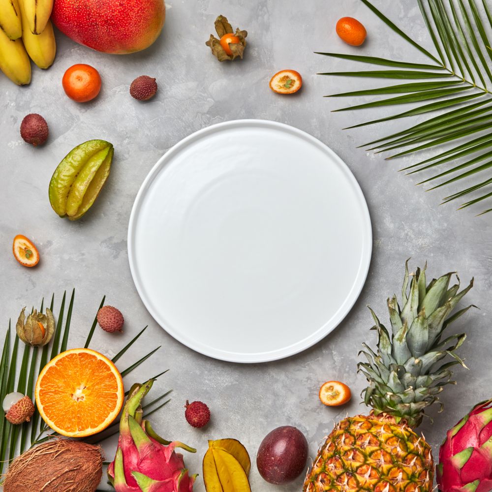 Composition from an empty white plate of green palm leaves, pineapple, mango,carambola, lychee, mandarin, and passion fruit on a gray marble table with a copy space. Flat lay. Palm leaves, empty white plate and a set of different tropical fruits mango, passion fruit,banana, coconut, physalis, pineapple on a gray concrete background with space for text. Flat lay