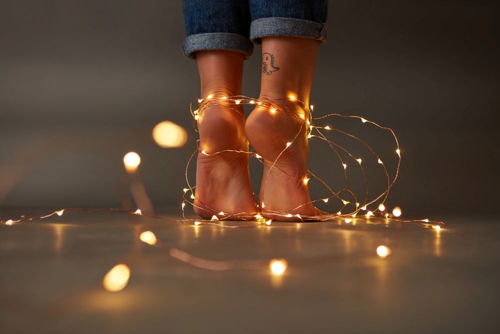 Beautiful female feet with yellow Christmas garlands on the floor around a dark background with space for text. Celebration. Composition of bright christmas lights decorating the feet of a young girl on a dark background