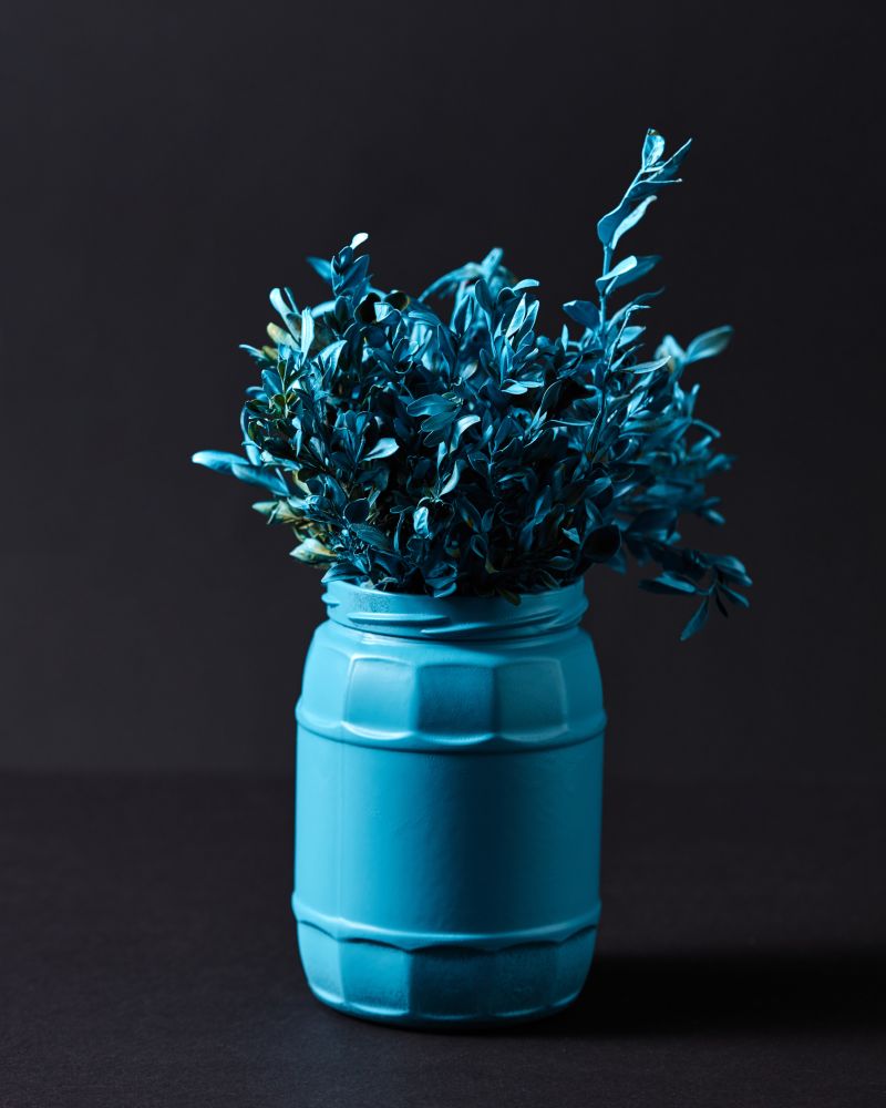 Painted blue branch of little leaf in a blue jar on a black background with copy space.. Blue vase with bouquet of twigs with blue painted leaves on a black background.