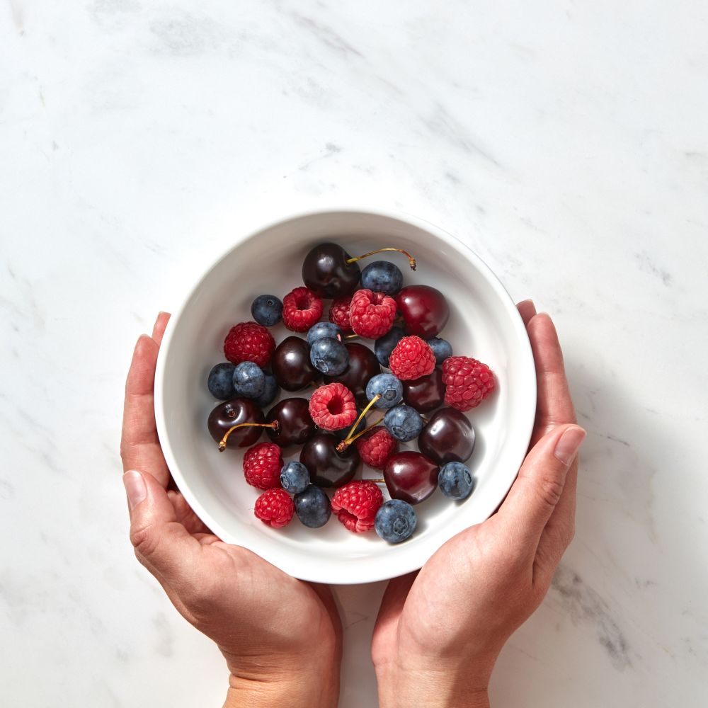Natural organic fruits, berries in a plate on a gray stone background. Concept of healthy food with copy space. Flat lay.. Women&rsquo;s hands hold a plate with ripe sweet berries - cherry, raspberry, blueberries on a gray stone background.