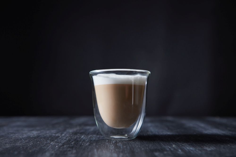 Freshly made cappuccino with foam in a glass cup on a wooden table around a dark background with space for text.. A glass cup of freshly made cappuccino presented on a black wooden table with copy space.