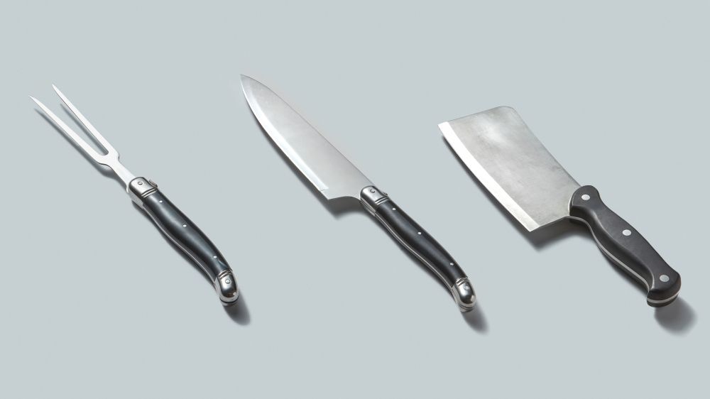 Meat cleaver, fork and knife from steel for cut fresh natural meat with shadows on a gray background, copy space. Steel kitchenware for cutting meat isolated on a gray background with shadows, place for text.