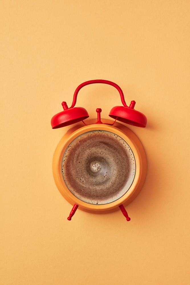 Vintage yellow alarm clock with red bells and cup of coffee on an yellow pastel background, place for text. Concept of morning coffee. Top view.. Retro style yellow alarm clock with red bells and fresh coffee instead of dial-plate on an yellow background place for text. Flat lay.